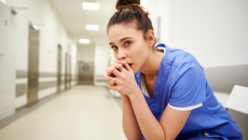 Eine Krankenschwester in blauer Arbeitskleidung sitzt nachdenklich auf einer Bank im Krankenhausflur und hält ihre Hände vor dem Gesicht.