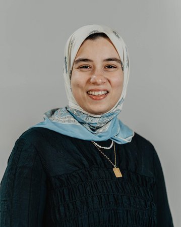 A young woman with a headscarf and a smiling face is wearing a black blouse and a blue scarf. The background is neutral.