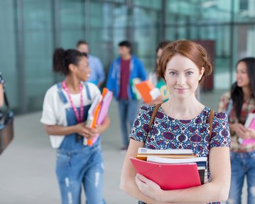 A student is holding several books and folders in her hands while other students in the background are talking and moving around.
