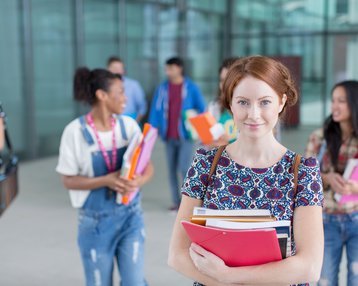 Eine Studentin hält mehrere Bücher und Ordner in den Händen, während andere Schüler im Hintergrund miteinander sprechen und sich bewegen.