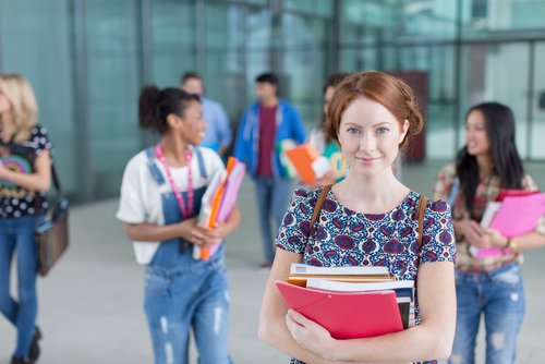 A student is holding several books and folders in her hands while other students in the background are talking and moving around.