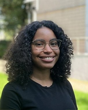 Eine Person mit lockigem, schwarzem Haar und Brille lächelt in die Kamera, im Hintergrund eine grüne Wiese und ein Gebäude.