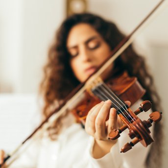 Eine Frau mit lockigem Haar spielt Geige in einem hellen Raum, während im Hintergrund eine Gitarre sichtbar ist. Sie konzentriert sich auf das Spiel und hält den Bogen in der rechten Hand.