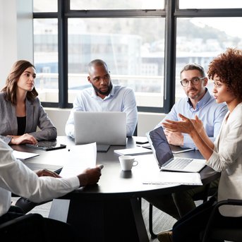 A group of five people is discussing in a meeting room. Some are using laptops while one person is speaking and the others are listening.