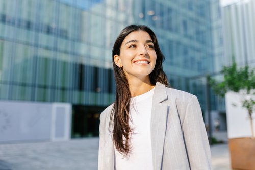 Junge Frau mit langen, dunklen Haaren und einem grauen Blazer lächelt vor einem modernen Gebäude mit Glasfassade.