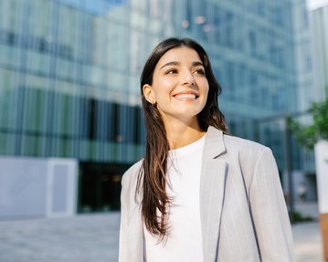 Junge Frau mit langen, dunklen Haaren und einem grauen Blazer lächelt vor einem modernen Gebäude mit Glasfassade.