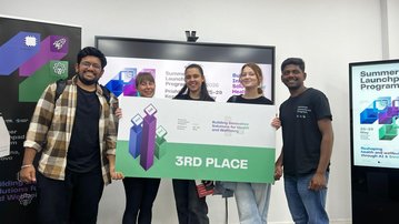 Fünf Personen stehen zusammen und halten ein Schild mit der Aufschrift "3. Platz" in einem Raum mit Bildschirmen im Hintergrund.