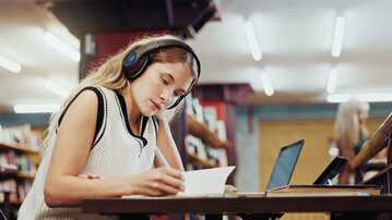Eine junge Frau mit Kopfhörern sitzt an einem Tisch in einer Bibliothek und schreibt in ein Notizbuch, während ein Laptop neben ihr steht.