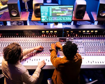 Zwei Personen arbeiten an einem Mischpult in einem Tonstudio. Einer dreht Regler, während der andere an einem Computerbildschirm mit Musiksoftware arbeitet. Lautsprecher sind im Hintergrund sichtbar.