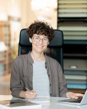 Eine Person mit lockigem Haar und Brille sitzt an einem Schreibtisch, arbeitet am Laptop und lächelt. Im Hintergrund sind Farbproben sichtbar.