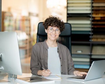 Eine Person mit lockigem Haar und Brille sitzt an einem Schreibtisch, arbeitet am Laptop und lächelt. Im Hintergrund sind Farbproben sichtbar.