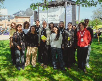 A group of people standing in front of a festival banner, surrounded by green spaces and trees, smiling at the camera.