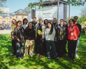 A group of people standing in front of a festival banner, surrounded by green spaces and trees, smiling at the camera.