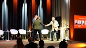 Drei Männer stehen auf einer Bühne und schütteln sich die Hände. Im Hintergrund sind leuchtende Vorhänge und Stühle zu sehen. Auf der rechten Seite ist ein Bildschirm mit Informationen zu den Panelteilnehmern.