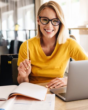 Frau mit Brille und gelben Pullover sitzt an ihrem Schreibtisch und lächelt in die Kamera
