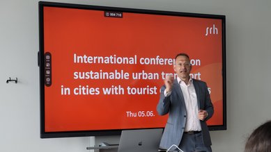 Ein Mann steht an einem Tisch und spricht während einer Präsentation. Im Hintergrund ist ein Bildschirm mit dem Titel "Internationale Konferenz über nachhaltigen urbanen Verkehr in Städten mit Touristenattraktionen" zu sehen. Das Datum ist der 5. Juni.