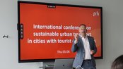 Ein Mann steht an einem Tisch und spricht während einer Präsentation. Im Hintergrund ist ein Bildschirm mit dem Titel "Internationale Konferenz über nachhaltigen urbanen Verkehr in Städten mit Touristenattraktionen" zu sehen. Das Datum ist der 5. Juni.