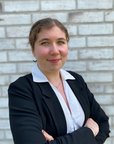 Eine junge Frau in einem schwarzen Blazer steht vor einer weißen Ziegelwand. Sie hat die Arme verschränkt und lächelt leicht, während sie in die Kamera schaut.