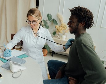 Eine Ärztin untersucht einen Patienten mit einem Stethoskop, während sie Notizen macht. Der Patient sitzt entspannt am Tisch, umgeben von Pflanzen und medizinischen Geräten.
