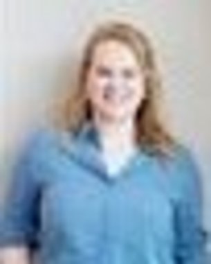 Eine lächelnde Frau mit blonden Haaren trägt ein blaues Hemd und steht vor einer neutralen Wand.