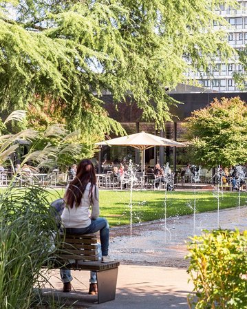 People are sitting on benches in a park with a fountain and trees. In the background, cafés and other visitors can be seen.
