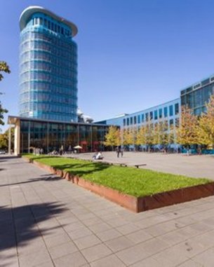 Moderne Architektur mit einem hohen Glasgebäude und einem begrünten Platz. Bäume und Menschen sind im Freien sichtbar, bei klarem Himmel und sonnigem Wetter.