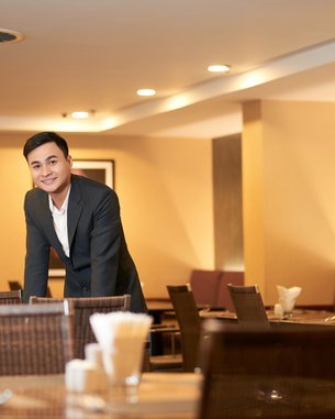 Ein Mann in einem Anzug steht an einem Tisch in einem Restaurant. Die Umgebung ist modern und gut beleuchtet. Im Hintergrund ist eine Person mit einem traditionellen Hut zu sehen.