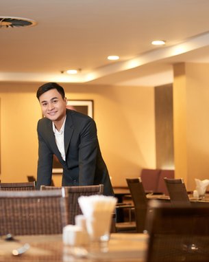 Ein Mann in einem Anzug steht an einem Tisch in einem Restaurant. Die Umgebung ist modern und gut beleuchtet. Im Hintergrund ist eine Person mit einem traditionellen Hut zu sehen.