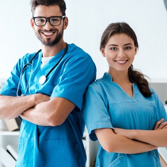 Zwei medizinische Fachkräfte in blauen Kitteln stehen lächelnd mit verschränkten Armen vor einem Regal.