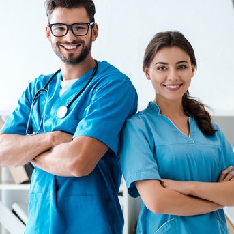 Two medical professionals in blue coats are standing with their arms crossed and smiling in front of a shelf.
