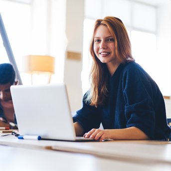 Eine lächelnde Frau sitzt an einem Tisch mit einem Laptop, während im Hintergrund zwei Personen an einem Projekt arbeiten. Helle, moderne Umgebung mit natürlichem Licht.