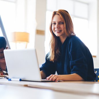 Eine lächelnde Frau sitzt an einem Tisch mit einem Laptop, während im Hintergrund zwei Personen an einem Projekt arbeiten. Helle, moderne Umgebung mit natürlichem Licht.