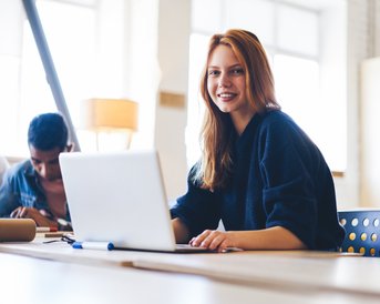 A smiling woman sits at a table with a laptop, while in the background, two people are working on a project. A bright, modern environment with natural light.