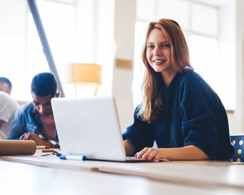 Eine lächelnde Frau sitzt an einem Tisch mit einem Laptop, während im Hintergrund zwei Personen an einem Projekt arbeiten. Helle, moderne Umgebung mit natürlichem Licht.