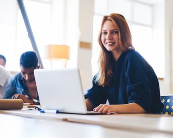 A smiling woman sits at a table with a laptop, while in the background, two people are working on a project. A bright, modern environment with natural light.