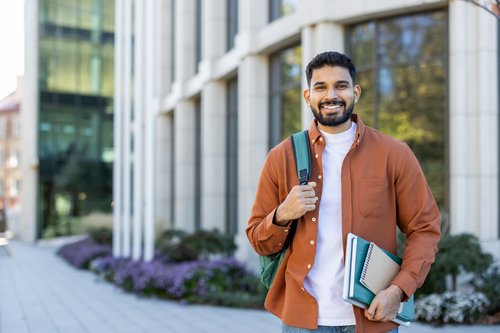 A young man with a beard is wearing an orange jacket and carrying a backpack. He is holding books and standing in front of a modern building.