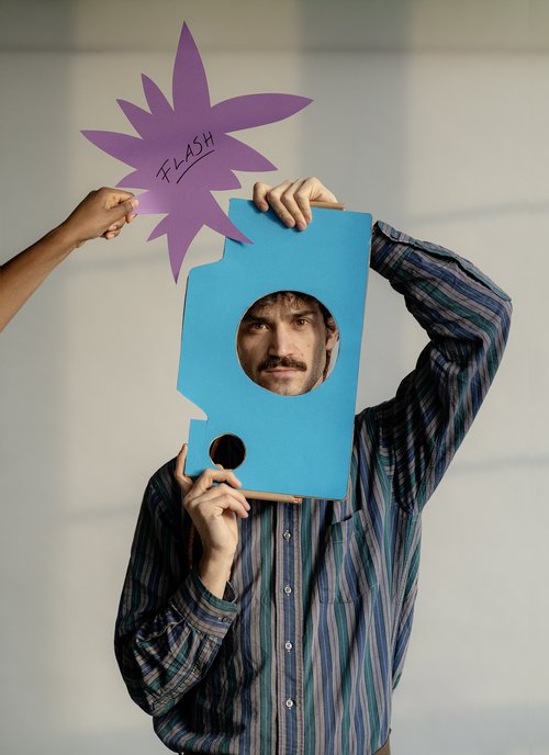 Ein Mann hält ein blaues Pappstück mit einem Loch und posiert, während eine Hand ein lila Schild mit der Aufschrift "FLASH" hält.