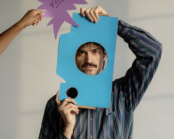 A man is holding a blue piece of cardboard with a hole in it and posing, while a hand holds a purple sign that says 'FLASH.