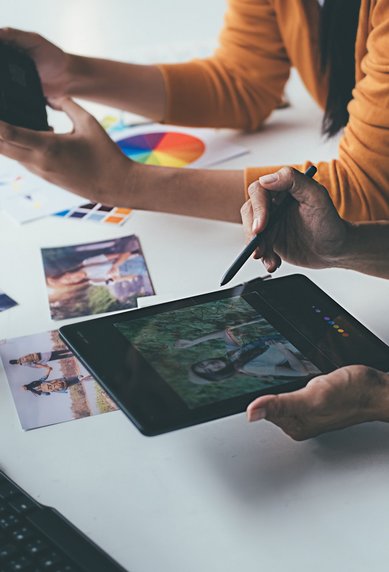 Two people are working at a table; one is using a tablet for photo editing while the other is holding a camera. Color prints are scattered around.