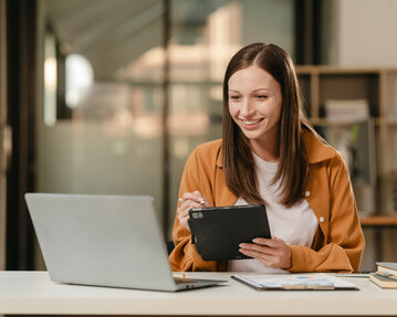A smiling woman is sitting at a table, using a tablet and looking at a laptop screen. Books are lying next to her.