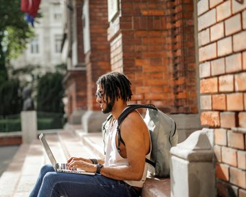 Ein Mann mit Dreadlocks sitzt auf einer Treppe und arbeitet an einem Laptop. Er trägt einen Rucksack und hat ein lässiges Outfit an. Im Hintergrund sind Bäume und eine Ziegelmauer sichtbar.