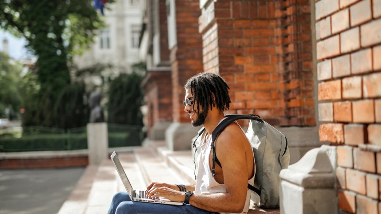 Ein Mann mit Dreadlocks sitzt auf einer Treppe und arbeitet an einem Laptop. Er trägt einen Rucksack und hat ein lässiges Outfit an. Im Hintergrund sind Bäume und eine Ziegelmauer sichtbar.