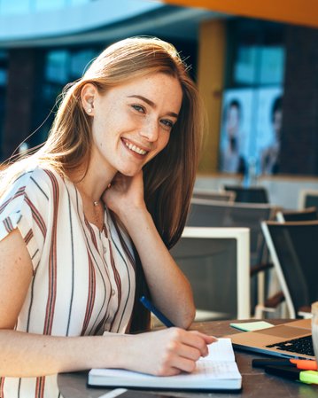 Eine junge Frau mit langen, roten Haaren sitzt an einem Tisch, schreibt in ein Notizbuch und lächelt. Vor ihr steht ein Laptop und ein Glas Getränk.