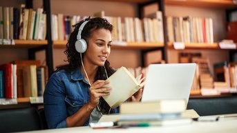 A woman with headphones is reading a book while working on a laptop. In the background, bookshelves are visible.