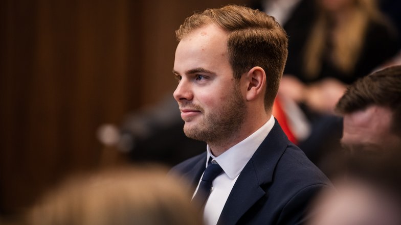 Ein junger Mann mit kurzen, blonden Haaren und Anzug sitzt aufmerksam in einer Versammlung.