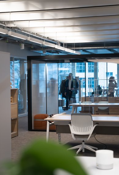 A modern office with bright furniture, large windows, and plants. Two people enter the room, while a table and chairs are visible in the foreground.