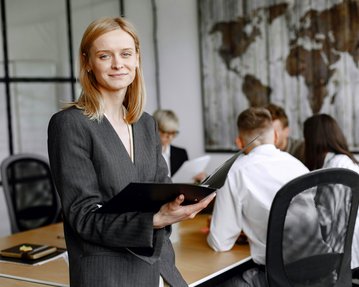 Eine Frau in einem grauen Anzug hält ein Notizbuch und lächelt, während im Hintergrund eine Gruppe von Personen in einem Meeting sitzt.