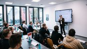 Ein Dozent präsentiert vor einer Gruppe von Studenten in einem modernen Klassenraum, während ein Bildschirm Informationen anzeigt.