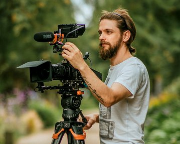Ein Mann mit Bart stellt eine Kamera auf einem Stativ ein, während er in einem Garten steht. Die Kamera hat ein Mikrofon montiert.