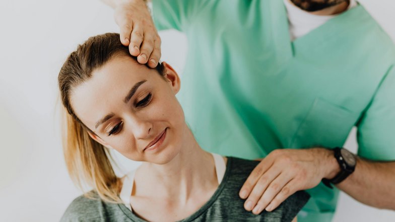 Eine Frau erhält eine Nackenmassage von einem männlichen Therapeuten. Der Therapeut hat seine Hand auf ihren Kopf gelegt, während sie entspannt aussieht. Beide tragen bequeme Kleidung.